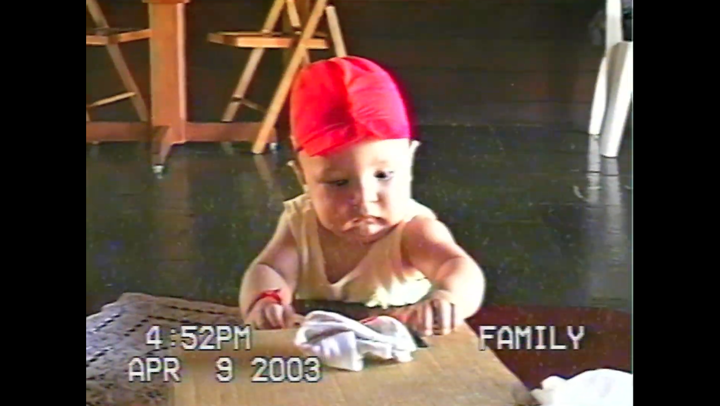 Bebê usando um gorro vermelho, vestindo uma regata clara, sentado próximo a uma mesa com um pano branco. A imagem tem um estilo de vídeo caseiro antigo, com marcações de data e hora mostrando 4:52 PM, 9 de abril de 2003, e a palavra 'FAMILY' no canto inferior direito.